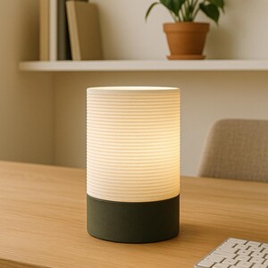 Puede incluir: Una l&aacute;mpara de mesa cil&iacute;ndrica blanca y verde con una pantalla blanca acanalada y una base verde oscuro. La l&aacute;mpara est&aacute; sobre una mesa de madera con un teclado.