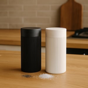 Op de afbeelding: Twee cilindrische zout- en peperstrooiers. De ene is zwart, de andere wit. Beide hebben een geribbeld ontwerp op de behuizing en een gladde, platte bovenkant. Zout en peper zijn op het houten oppervlak gestrooid.