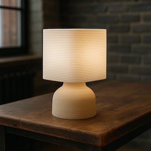 May include: A cream-colored table lamp with a square lampshade featuring horizontal lines. The lamp has a rounded base and sits on a dark wooden table. The background includes a brick wall and a window.