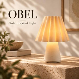 Op de afbeelding: Een cr&egrave;mekleurige tafellamp met een geplooide, uitlopende kap en een conische voet. De lamp geeft een warm, zacht licht. De tekst OBEL en Soft pleated light is zichtbaar in de linkerbovenhoek van de afbeelding tegen een neutrale achtergrond.