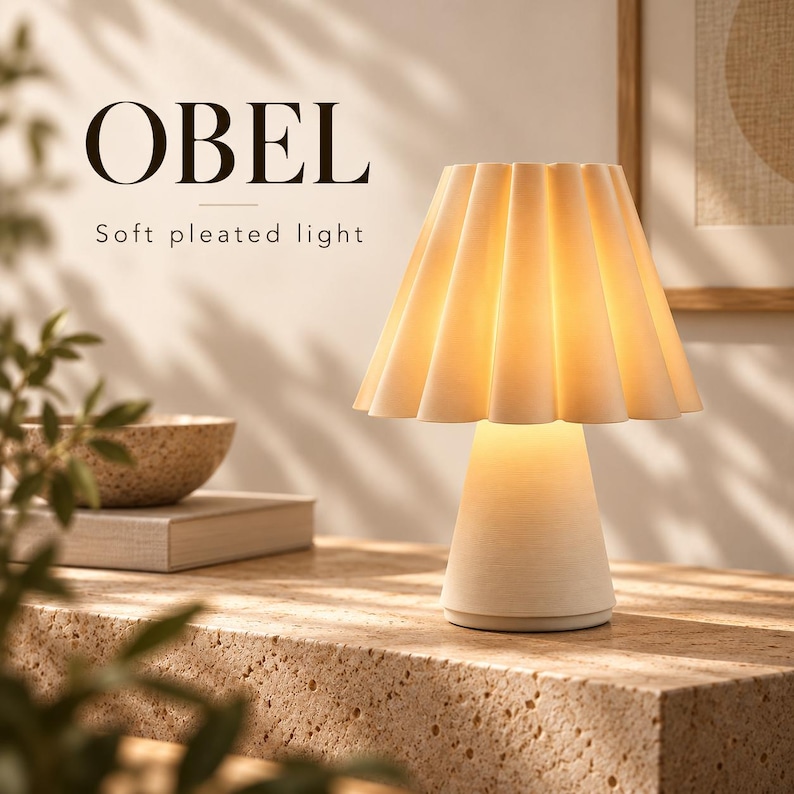 Op de afbeelding: Een cr&egrave;mekleurige tafellamp met een geplooide, uitlopende kap en een conische voet. De lamp geeft een warm, zacht licht. De tekst OBEL en Soft pleated light is zichtbaar in de linkerbovenhoek van de afbeelding tegen een neutrale achtergrond.