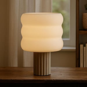 May include: A modern table lamp with a cream-colored, rounded shade and a ribbed, cylindrical base. The lamp sits on a dark wooden surface, with a neutral background. The lamp's design is minimalist and contemporary, suitable for home decor.