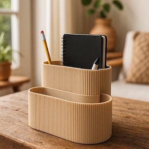 Puede incluir: Un organizador de escritorio beige con diseño acanalado, que contiene un lápiz amarillo, un cuaderno negro y un rotulador blanco. El organizador está sobre una mesa de madera, ideal para un espacio de trabajo.