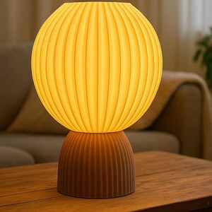 Puede incluir: Una l&aacute;mpara de mesa con una pantalla esf&eacute;rica acanalada que emite un c&aacute;lido resplandor amarillo, apoyada sobre una base m&aacute;s peque&ntilde;a y a juego con la misma textura acanalada en un tono marr&oacute;n oscuro y apagado. El dise&ntilde;o consta de dos piezas apiladas.
