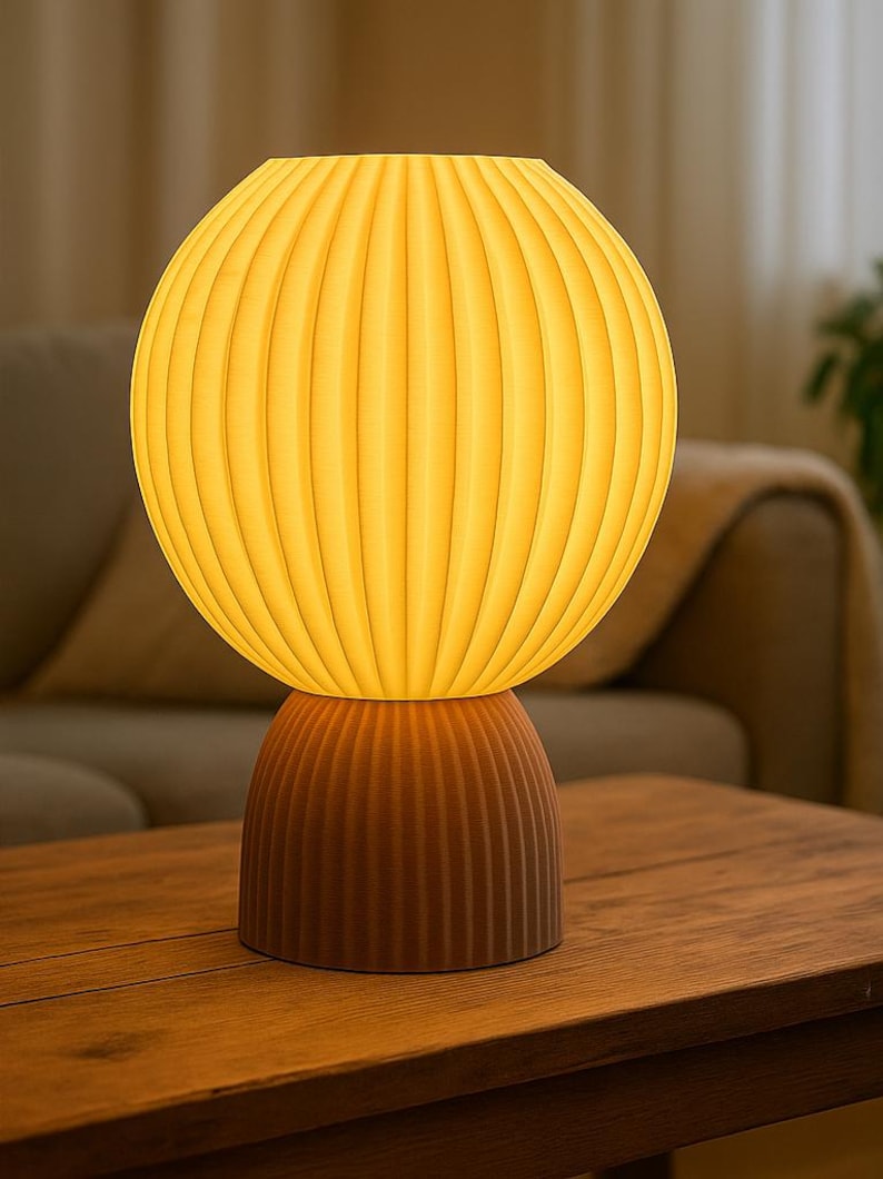 Puede incluir: Una l&aacute;mpara de mesa con una pantalla esf&eacute;rica acanalada que emite un c&aacute;lido resplandor amarillo, apoyada sobre una base m&aacute;s peque&ntilde;a y a juego con la misma textura acanalada en un tono marr&oacute;n oscuro y apagado. El dise&ntilde;o consta de dos piezas apiladas.