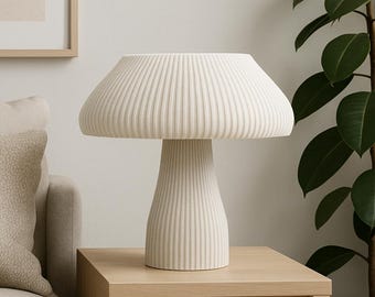 May include: A cream-colored mushroom-shaped table lamp with a ribbed lampshade and a conical base. The lamp sits on a light-colored wooden side table. A framed picture and a plant are in the background.