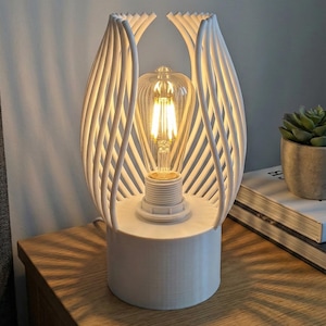 Może przedstawiać: Biała, nowoczesna lampa stołowa o unikalnym, ażurowym designie. Lampa posiada cylindryczną podstawę i klosz w kształcie klatki, złożony z pionowych, zakrzywionych linii. Wewnątrz widoczna jest żarówka w stylu vintage. Lampa stoi na drewnianej powierzchni.