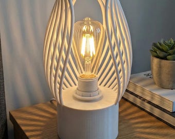 May include: White, modern table lamp with a unique, openwork design. The lamp features a cylindrical base and a cage-like shade composed of vertical, curved lines. A vintage-style light bulb is visible inside. The lamp is on a wooden surface.