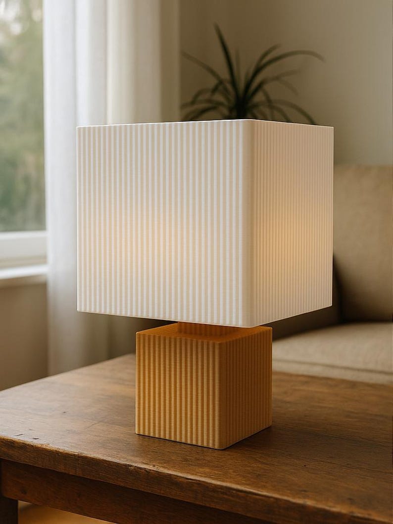 Puede incluir: Una l&aacute;mpara de mesa moderna con una base y una pantalla en forma de cubo. La base es de color amarillo c&aacute;lido con ranuras verticales, y la pantalla es blanca con rayas verticales. La l&aacute;mpara est&aacute; sobre una mesa de madera oscura.