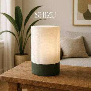 Puede incluir: Una l&aacute;mpara de mesa blanca y verde con una pantalla blanca texturizada y una base verde oscuro. La l&aacute;mpara est&aacute; encendida e ilumina la habitaci&oacute;n. La l&aacute;mpara est&aacute; sobre una mesa de madera. El texto "SHIZU" es visible sobre la l&aacute;mpara.