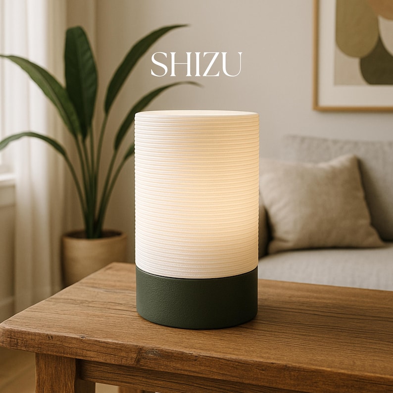 Puede incluir: Una l&aacute;mpara de mesa blanca y verde con una pantalla blanca texturizada y una base verde oscuro. La l&aacute;mpara est&aacute; encendida e ilumina la habitaci&oacute;n. La l&aacute;mpara est&aacute; sobre una mesa de madera. El texto "SHIZU" es visible sobre la l&aacute;mpara.