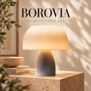 Op de afbeelding: Een tafellamp met een paddenstoelvormige, geribbelde witte kap en een taps toelopende, geribbelde grijze voet. De tekst BOROVIA Sculptural ribbed glow is boven de lamp zichtbaar. De lamp verspreidt een warm, zacht licht over de textuur.