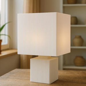 Puede incluir: L&aacute;mpara de mesa moderna de color crema con pantalla y base cuadradas. La l&aacute;mpara presenta detalles acanalados verticales y se encuentra sobre una mesa de madera. El dise&ntilde;o es minimalista y contempor&aacute;neo, adecuado para la decoraci&oacute;n del hogar. Medidas en cent&iacute;metros.