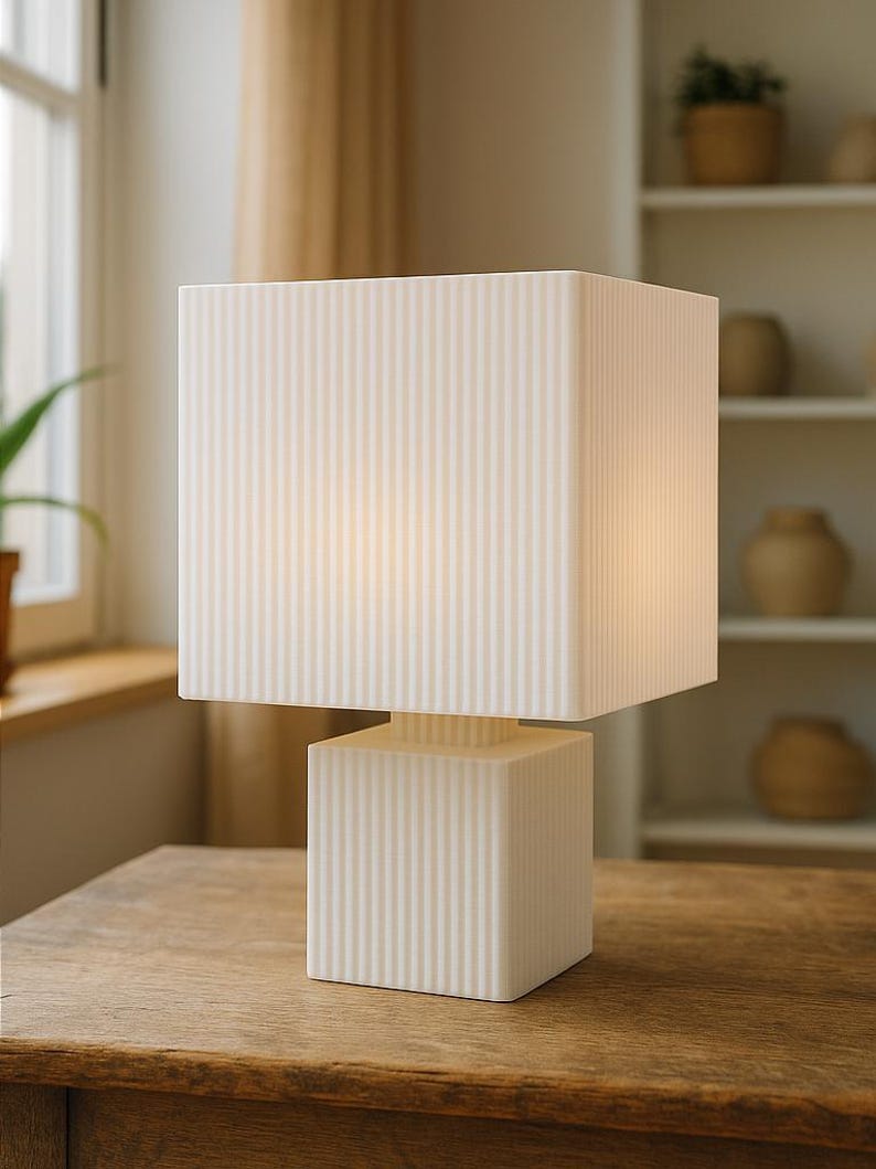 Puede incluir: L&aacute;mpara de mesa moderna de color crema con pantalla y base cuadradas. La l&aacute;mpara presenta detalles acanalados verticales y se encuentra sobre una mesa de madera. El dise&ntilde;o es minimalista y contempor&aacute;neo, adecuado para la decoraci&oacute;n del hogar. Medidas en cent&iacute;metros.