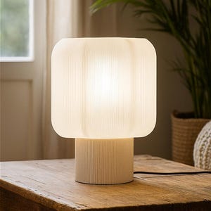 Könnte beinhalten: Eine cremefarbene Tischlampe mit geripptem Sockel und quadratischem, abgerundetem Schirm. Die Lampe ist beleuchtet und wirft ein sanftes Licht. Sie steht auf einem Holztisch, mit einem Fenster und Grün im Hintergrund.