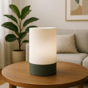 Puede incluir: Una l&aacute;mpara de mesa blanca y gris con una pantalla blanca texturizada y una base gris oscuro. La l&aacute;mpara est&aacute; sobre una mesa de madera.