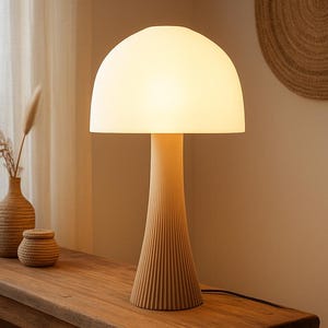Pode incluir: Uma luminária de mesa bege com uma cúpula branca brilhante em forma de cogumelo. A luminária tem uma base cônica canelada e um pescoço cilíndrico. A luminária está em uma mesa de madeira, com um vaso e uma pequena tigela decorativa.