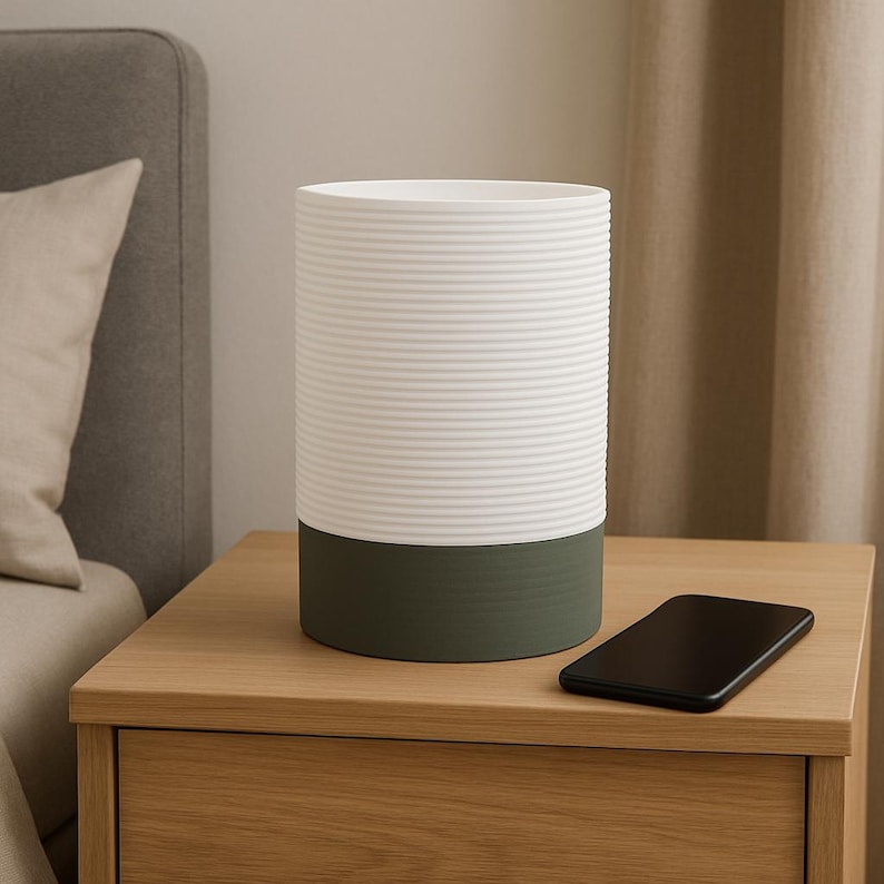 Puede incluir: Un jarr&oacute;n cil&iacute;ndrico de cer&aacute;mica blanco y verde con textura acanalada. El jarr&oacute;n est&aacute; sobre una mesita de noche de madera con un tel&eacute;fono m&oacute;vil en la superficie.