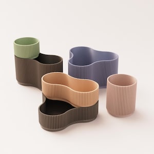 May include: A collection of modern, ribbed-textured containers in various shapes and colors. The set includes a light green cup, a wavy lavender container, a double-lobed tan and black container, and a pink cup. The containers are made of a solid material.