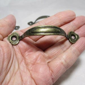 May include: Antique brass-toned drawer pulls. The pulls have a curved handle with decorative floral-shaped ends. The hardware is a warm, aged brass color, suggesting a vintage or classic aesthetic. Several pulls are visible.