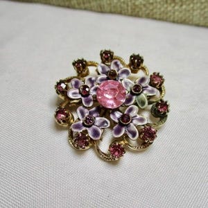 May include: A vintage floral brooch with a gold-tone metal base. The brooch features a central pink gemstone surrounded by purple and white enamel flowers and smaller pink gemstones. The design is intricate and detailed.