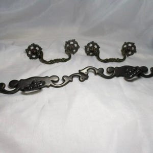 May include: A set of antique bronze-colored drawer pulls and handles. The set includes three smaller pulls with a floral design and a larger handle with a similar design. The hardware is ornate and likely for a cabinet or dresser.