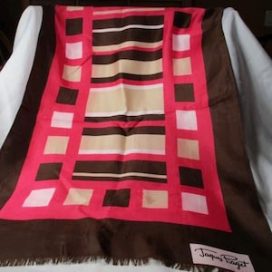 May include: A rectangular scarf with a geometric pattern in shades of brown, pink, beige, and white. The scarf has a fringed edge and a label that reads "Jacques Faget".