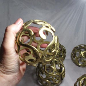May include: Ornate, gold-colored decorative spheres with openwork designs. The spheres have a detailed, swirling pattern and are likely made of metal. Several spheres are visible, with one held in a hand.