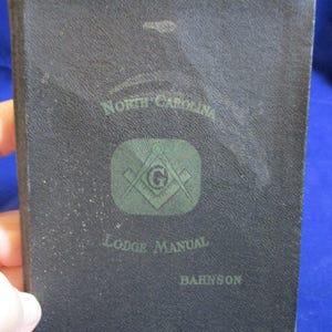 May include: A black hardcover book titled North Carolina Lodge Manual by Bahnson. The cover features a green square and compass emblem with a central letter G. The textured surface of the book displays the title and author in green lettering.