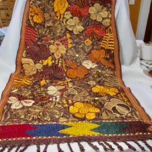 May include: A brown textile with embroidered floral patterns in shades of yellow, orange, and cream. The edges are trimmed with a woven border of red, blue, yellow, and green, and finished with a fringe.