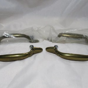 May include: Four brass-toned cabinet handles with mounting screws. Two are in plastic packaging, and two are visible in the foreground. The handles have a curved design and are likely for furniture or cabinetry.