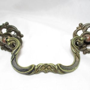 May include: Ornate antique bronze drawer pulls with a decorative floral design. The pulls have a curved handle and a detailed backplate. These vintage hardware pieces add a touch of elegance to any furniture.