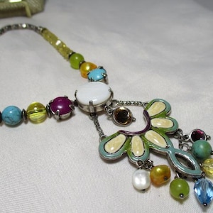May include: A colorful necklace with a silver chain and a pendant featuring a floral design. The necklace includes various beads in shades of yellow, turquoise, purple, and green, with a large white stone at the center.