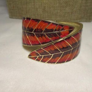May include: A gold-toned cuff bracelet with a leaf design. The leaves are in shades of red, orange, and brown, with gold outlines. The bracelet is on a white surface.