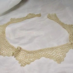 May include: A cream-colored, open-weave crochet collar with a scalloped edge. The collar is laid flat on a white surface. The delicate design suggests a vintage or handmade item, suitable for adding a touch of elegance to an outfit.