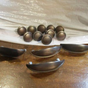 May include: A collection of cabinet hardware consisting of ten round, dark bronze-toned knobs and four matching crescent-shaped, semi-circular drawer pulls. Each piece features a smooth, matte finish with a consistent metallic dark brown color.