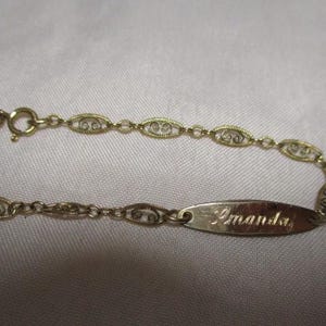 May include: A delicate gold-toned bracelet with an oval nameplate engraved with the name "Amanda". The bracelet features an ornate filigree design with oval links and a secure clasp.