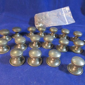 May include: A collection of twenty round cabinet knobs with a dark metallic finish. Each knob features a smooth, domed top and a flared base. A clear plastic bag containing small metal hardware components is positioned behind the knobs.