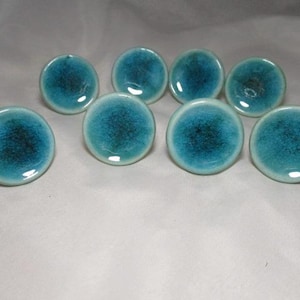 May include: Eight round ceramic cabinet knobs with a glossy finish. The knobs are turquoise with a dark blue, speckled center. The knobs are arranged in a scattered pattern on a light-colored surface.