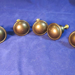 May include: Five round cabinet knobs with a smooth, dark bronze finish. Each knob features a circular, slightly domed face with a raised outer rim and a central mounting screw extending from the back, arranged in a horizontal row.
