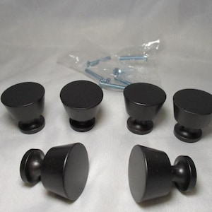 May include: A set of six black cabinet knobs with a conical design. The knobs are made of metal and have a matte finish. A small bag of screws is visible in the background, suggesting they are ready for installation.