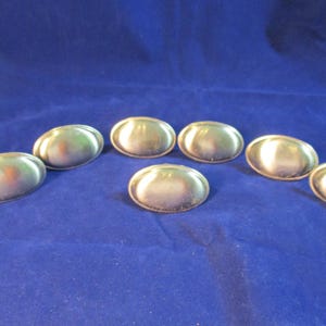 May include: Seven oval-shaped metal buttons with a smooth, reflective gold-tone finish. Each button features a slightly concave center and a raised, rounded rim, presenting a uniform appearance across the set of seven individual hardware pieces.