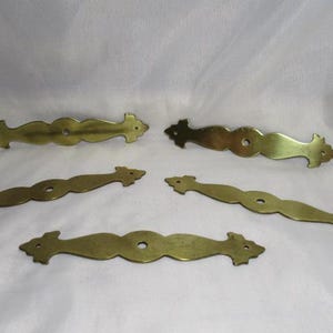 May include: Five antique brass drawer pulls with a decorative, symmetrical design. Each pull has a central oval section with a single hole, flanked by curved sections and decorative ends. The pulls are arranged on a white background.