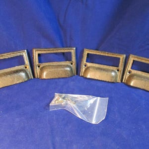 May include: Four rectangular metal drawer pulls with a dark, weathered finish and integrated label holders. Each pull features two circular screw holes for mounting. A small, clear plastic bag containing a set of metal screws is included.