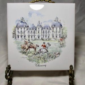 May include: White square tile with a watercolor painting of a large chateau with blue-domed roofs and riders on horseback. The tile has a gold-colored twisted metal stand. The word "Cheverny" is written at the bottom.