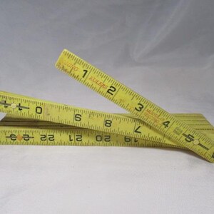 May include: A yellow folding ruler with black markings, showing measurements in inches. The ruler is partially open, displaying numbers and markings. The background is a plain white surface.