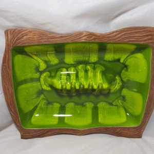 May include: A rectangular ashtray with a wavy, brown wooden frame. The interior is a vibrant green with a unique, abstract design. The ashtray is a decorative item.