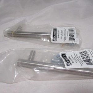 May include: Two stainless steel cabinet pulls in clear plastic packaging. Each pull has a cylindrical bar and a perpendicular handle. The packaging includes white labels with black text and barcodes.