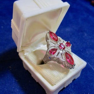 May include: A silver-toned ring featuring four marquise-cut red stones and one central oval-cut red stone, all surrounded by small clear pavé-set stones. The ring is displayed inside an open white plastic jewelry box with a cream-colored fabric lining.