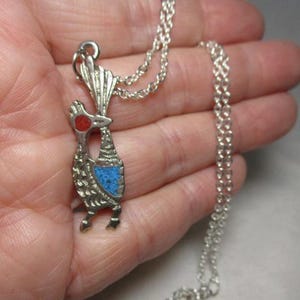 May include: A silver-toned pendant necklace featuring a stylized roadrunner design. The bird has a red eye and a blue mosaic-style chest. The necklace chain is also silver-toned.